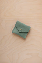 A small sage green suede jewelry pouch with a gold button fastening, resting on a light wood grain surface.