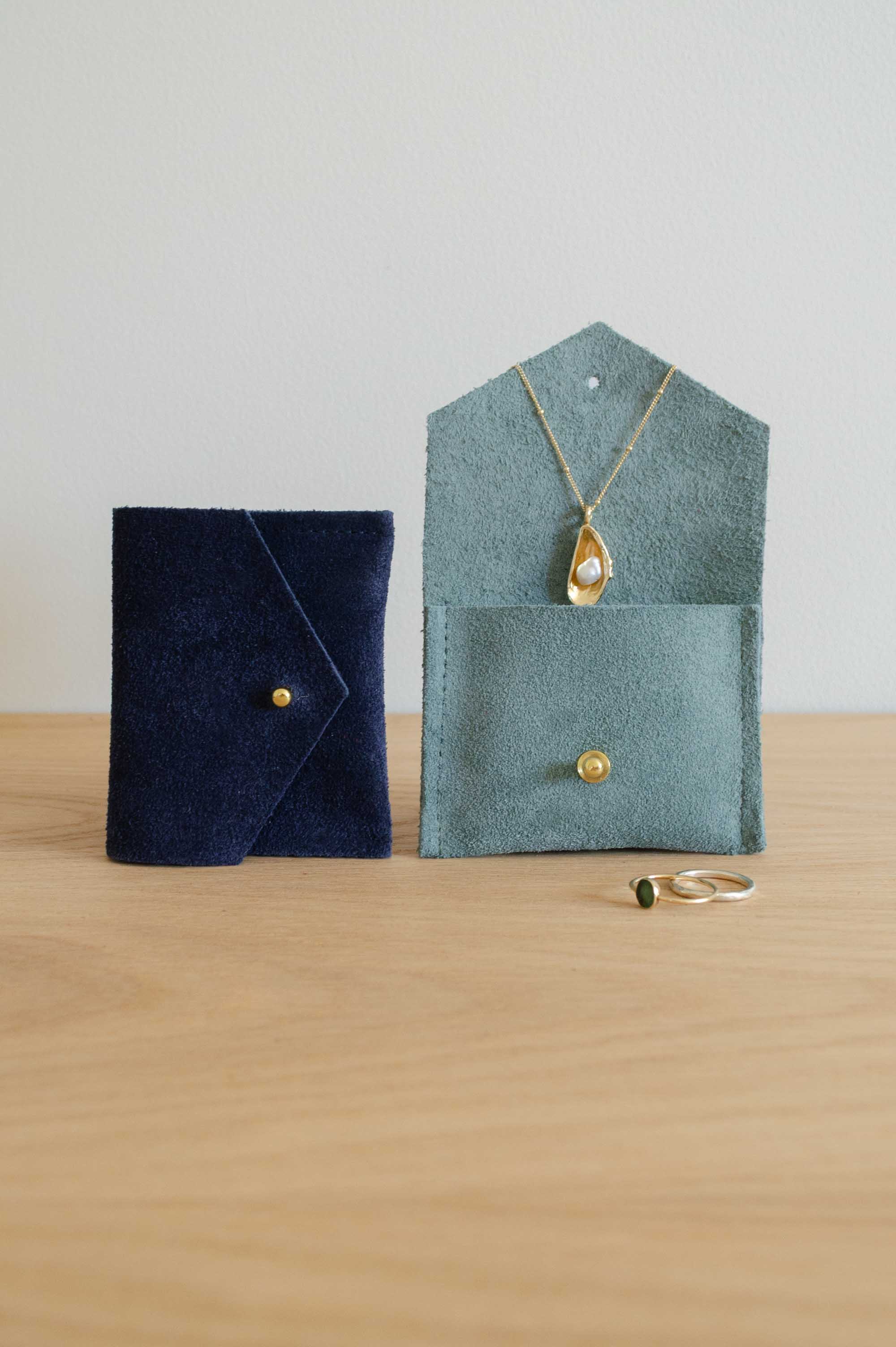 Two small suede jewelry pouches on a light wood surface against a plain light gray wall. One pouch is dark navy blue and the other is muted sage green, both with a button closure.