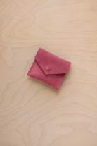 A small pink suede pouch with a flap closure and a tiny gold button, placed on a light wood grain surface.