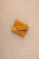 A small mustard yellow suede envelope-style pouch with a tiny gold button, placed on a light wood grain surface.