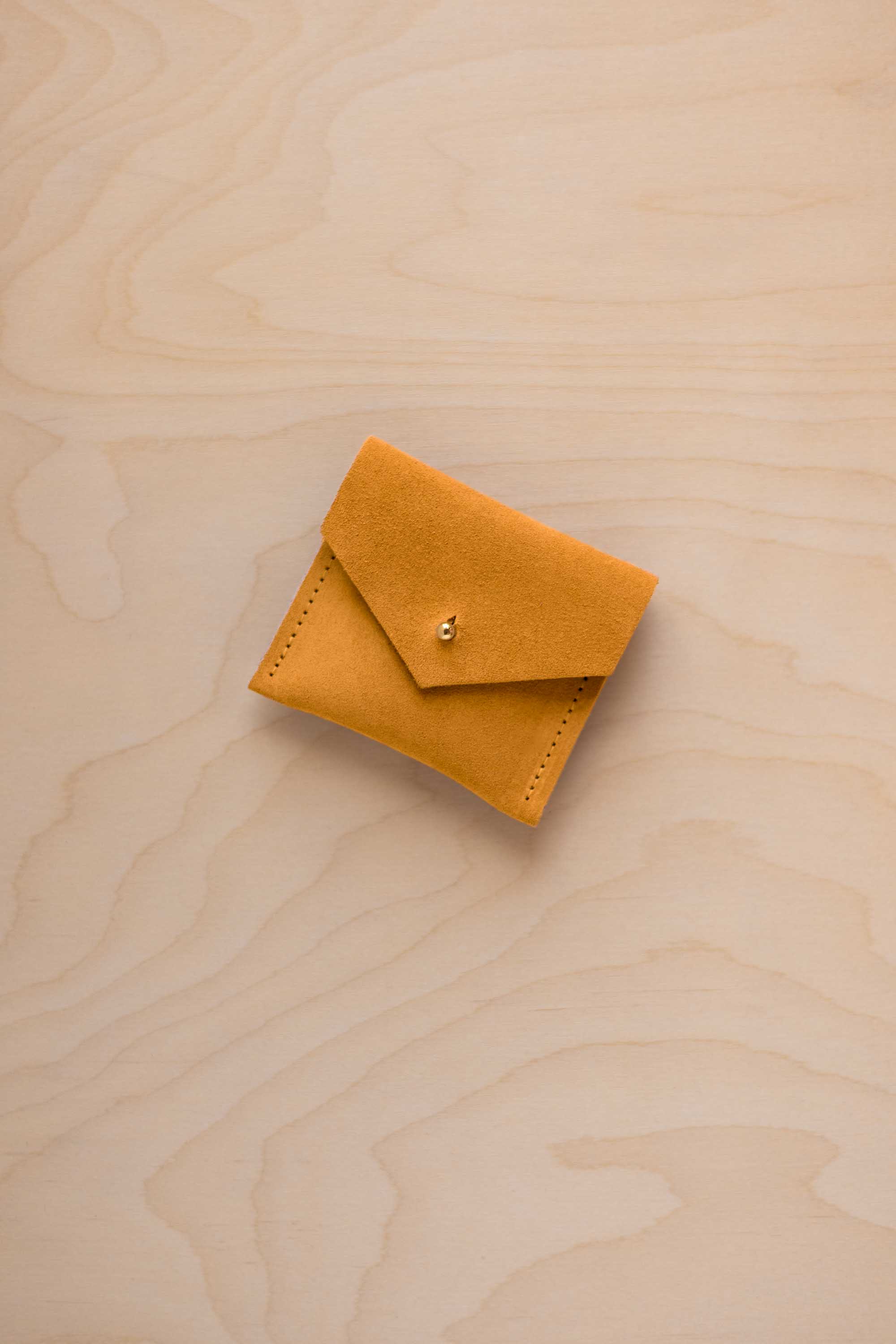 A small mustard yellow suede envelope-style pouch with a tiny gold button, placed on a light wood grain surface.