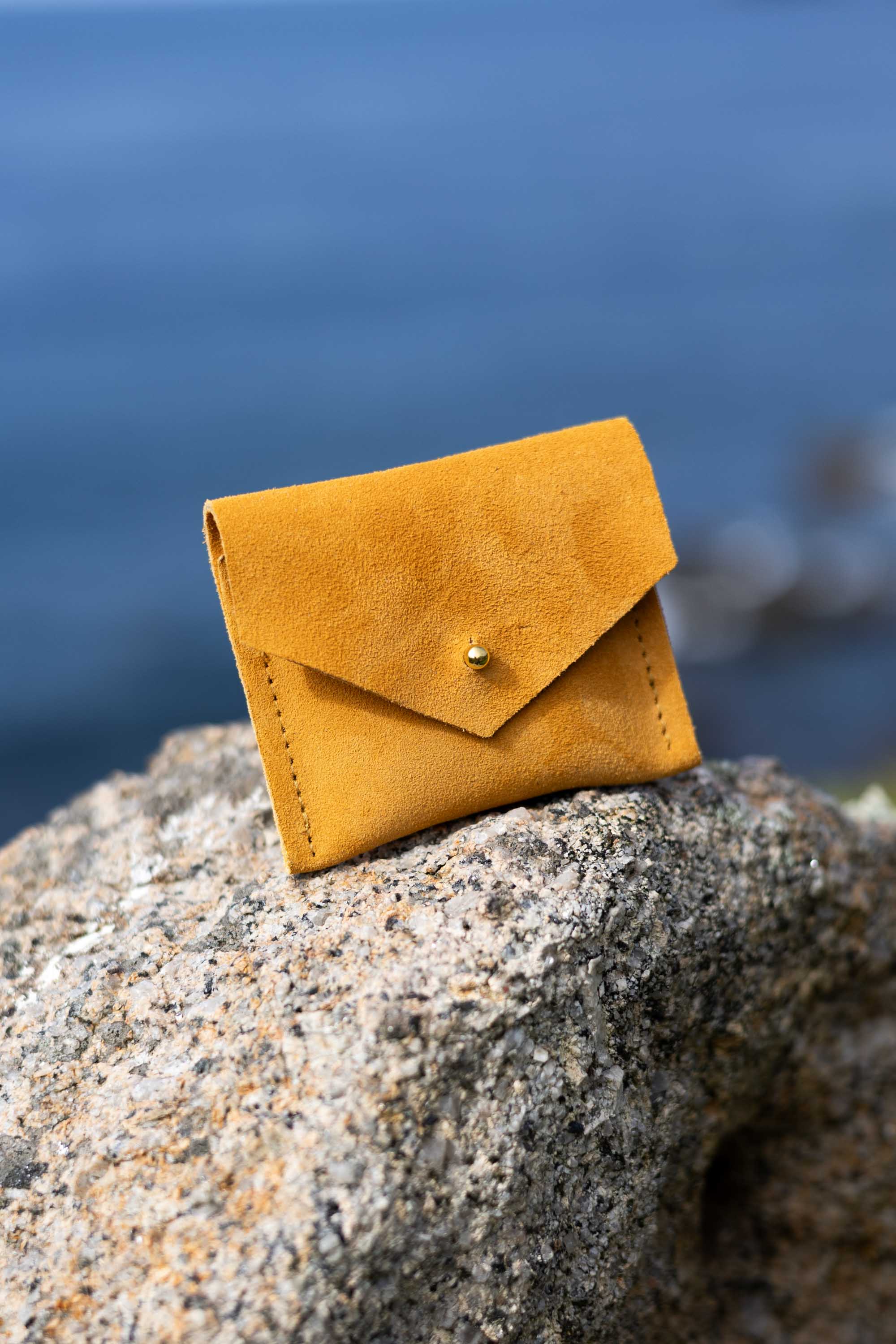 A small mustard yellow suede envelope-style card holder or wallet with a gold button, resting on a textured gray rock with a blurred blue water background.