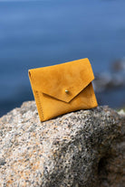 A small mustard yellow suede envelope-style card holder or wallet with a gold button, resting on a textured gray rock with a blurred blue water background.