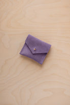 A small lavender purple suede jewelry pouch with a gold button, resting on a light wood grain background.