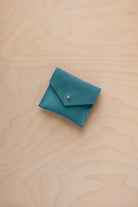 A small teal blue suede jewelry pouch with a gold button, resting on a light wood grain surface.