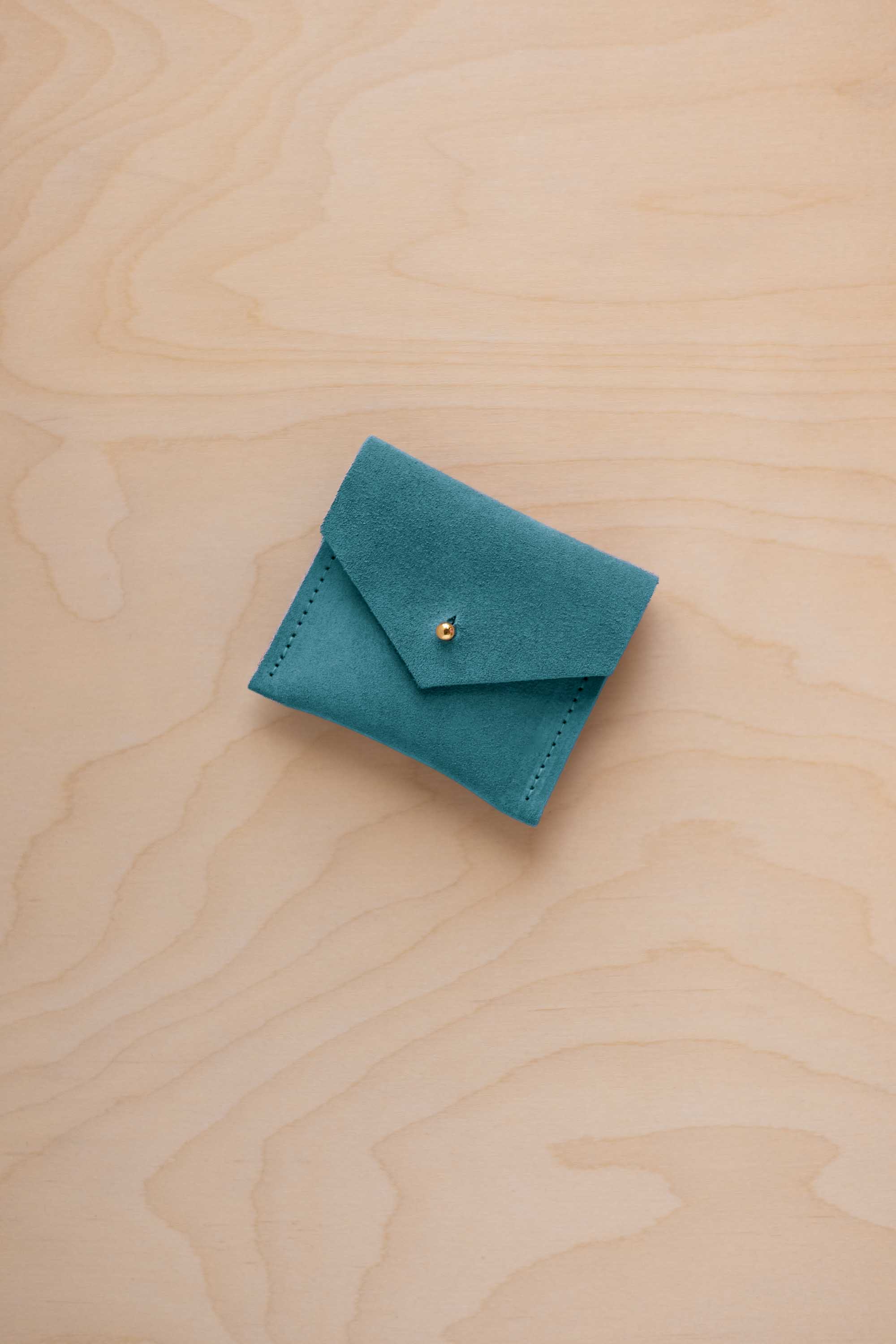 A small teal blue suede jewelry pouch with a gold button, resting on a light wood grain surface.