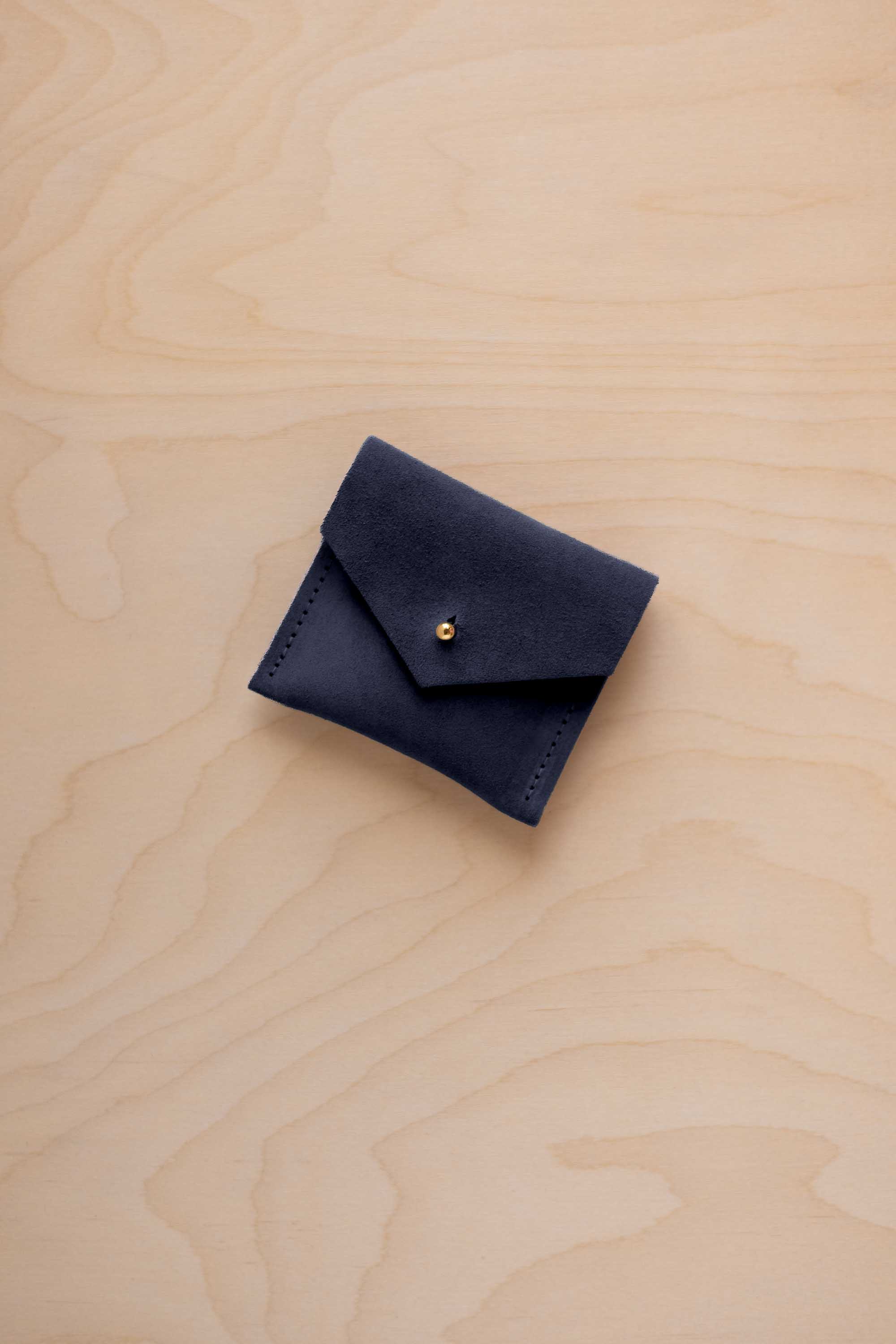 A small dark navy suede pouch with a flap closure and a gold-tone button, placed on a light wood grain surface.