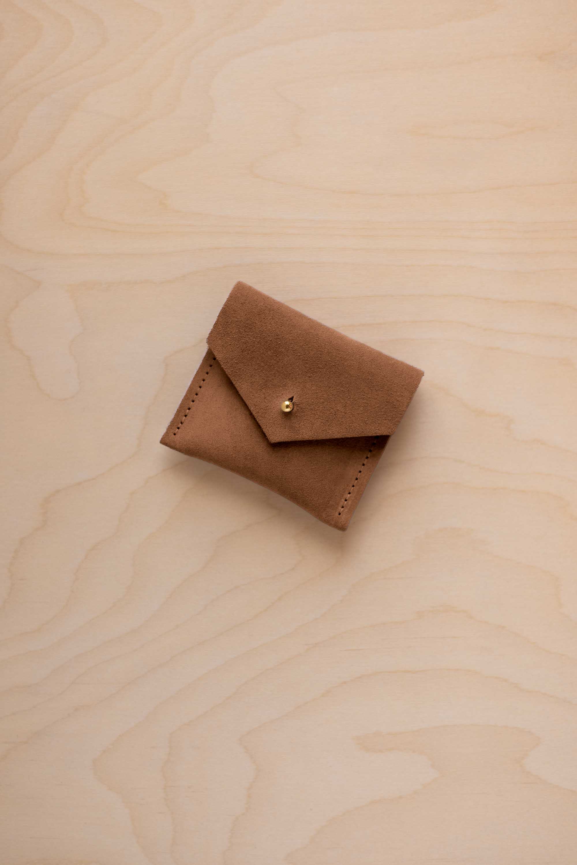 A small brown suede pouch with a flap closure and a gold button, resting on a light wood grain surface.