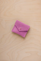 A small pink suede pouch with a flap closure and a tiny gold coloured button, placed on a light wood grain surface.