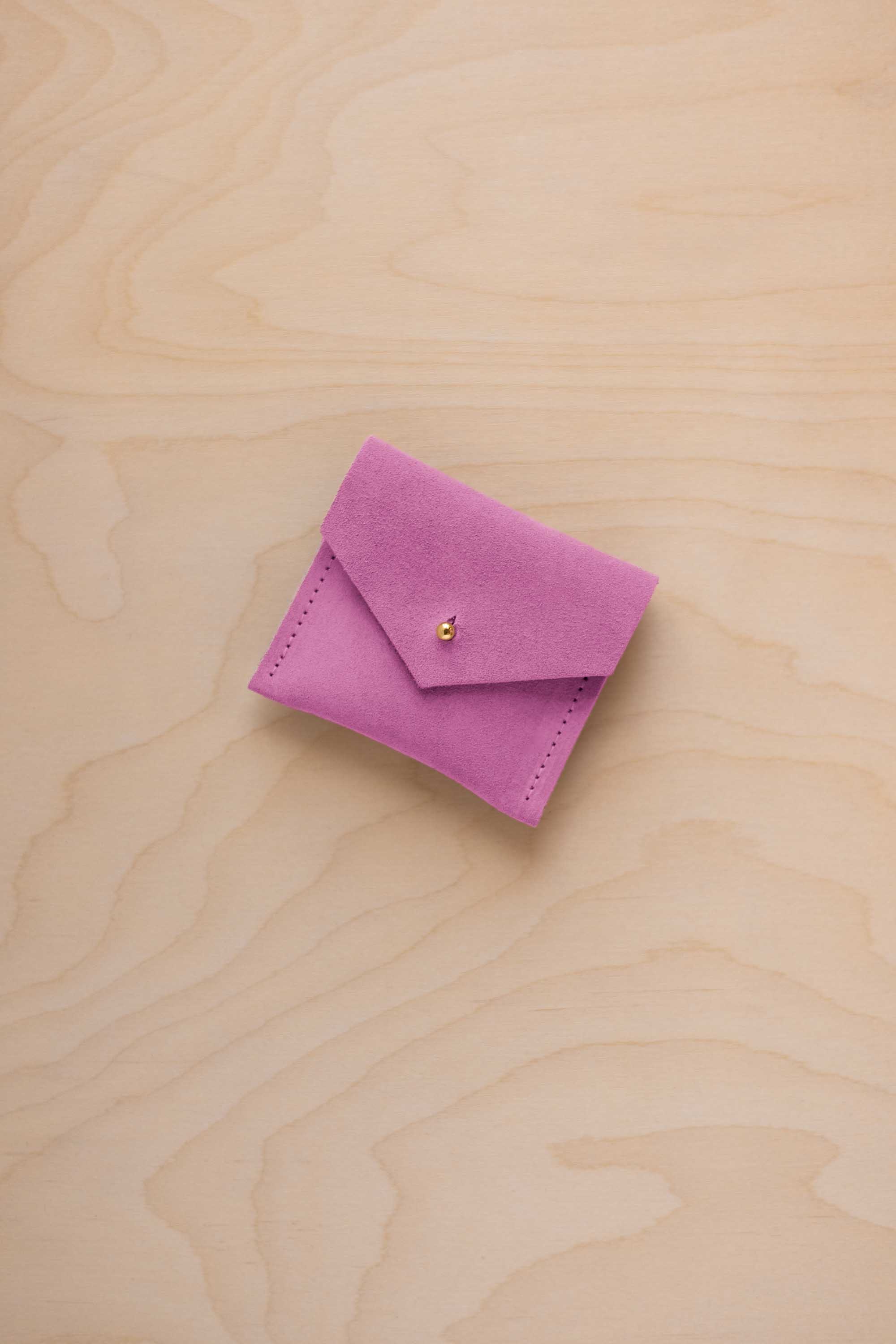A small pink suede pouch with a flap closure and a tiny gold coloured button, placed on a light wood grain surface.