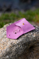 A small pink leather jewelry pouch or ring holder resting on a rough stone outdoors, with two rings placed inside and a small gold stud earring on the flap.