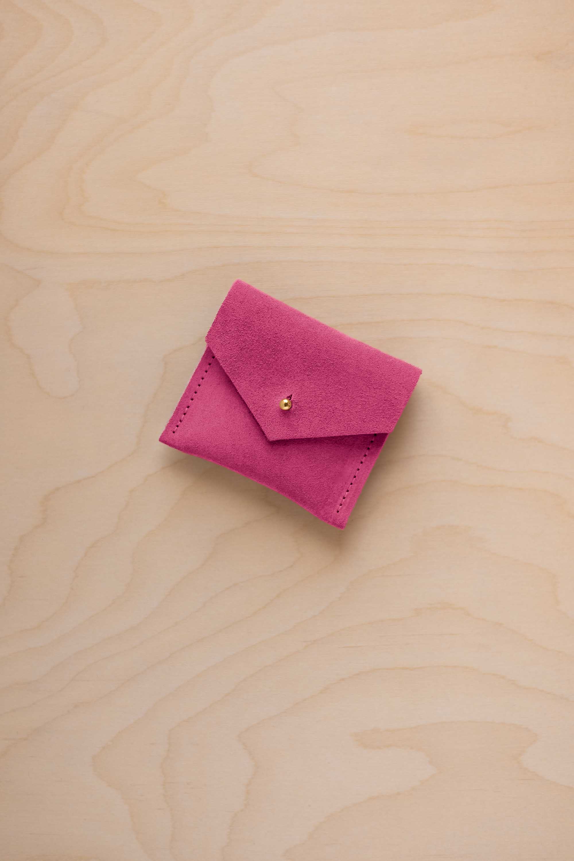 A small bright pink suede pouch with a flap closure and a tiny gold button, placed on a light wood grain surface.