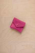 A small bright pink suede pouch with a flap closure and a tiny gold button, placed on a light wood grain surface.