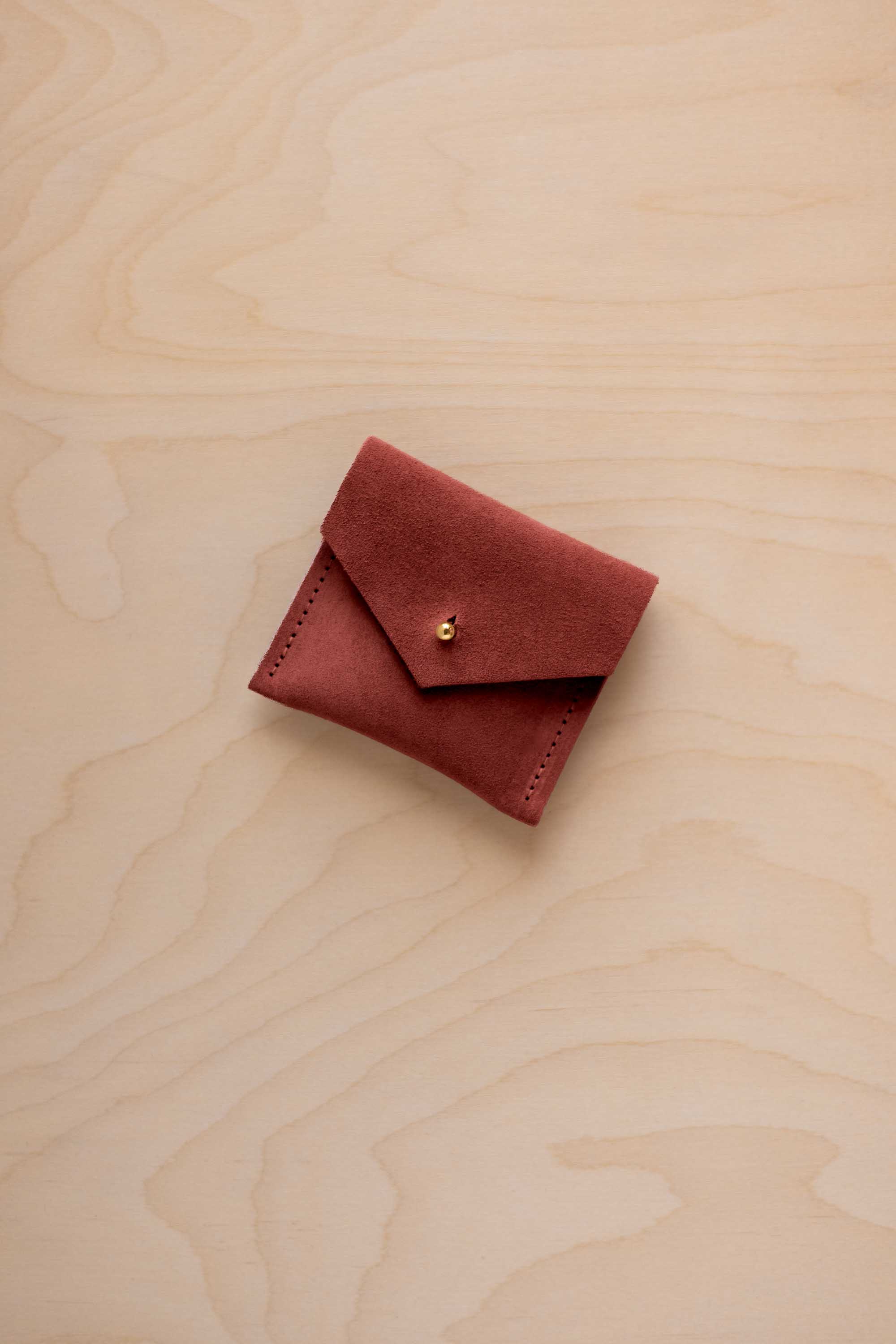 A small reddish-brown suede pouch with a flap closure and a gold button, placed on a light wood grain surface.
