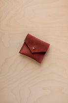 A small reddish-brown suede pouch with a flap closure and a gold button, placed on a light wood grain surface.