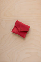 A small red suede pouch with a flap closure and a tiny gold button, placed on a light wood grain surface.