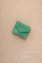 A small light emerald green suede pouch with a gold coloured button closure, placed on a light wood grain surface.