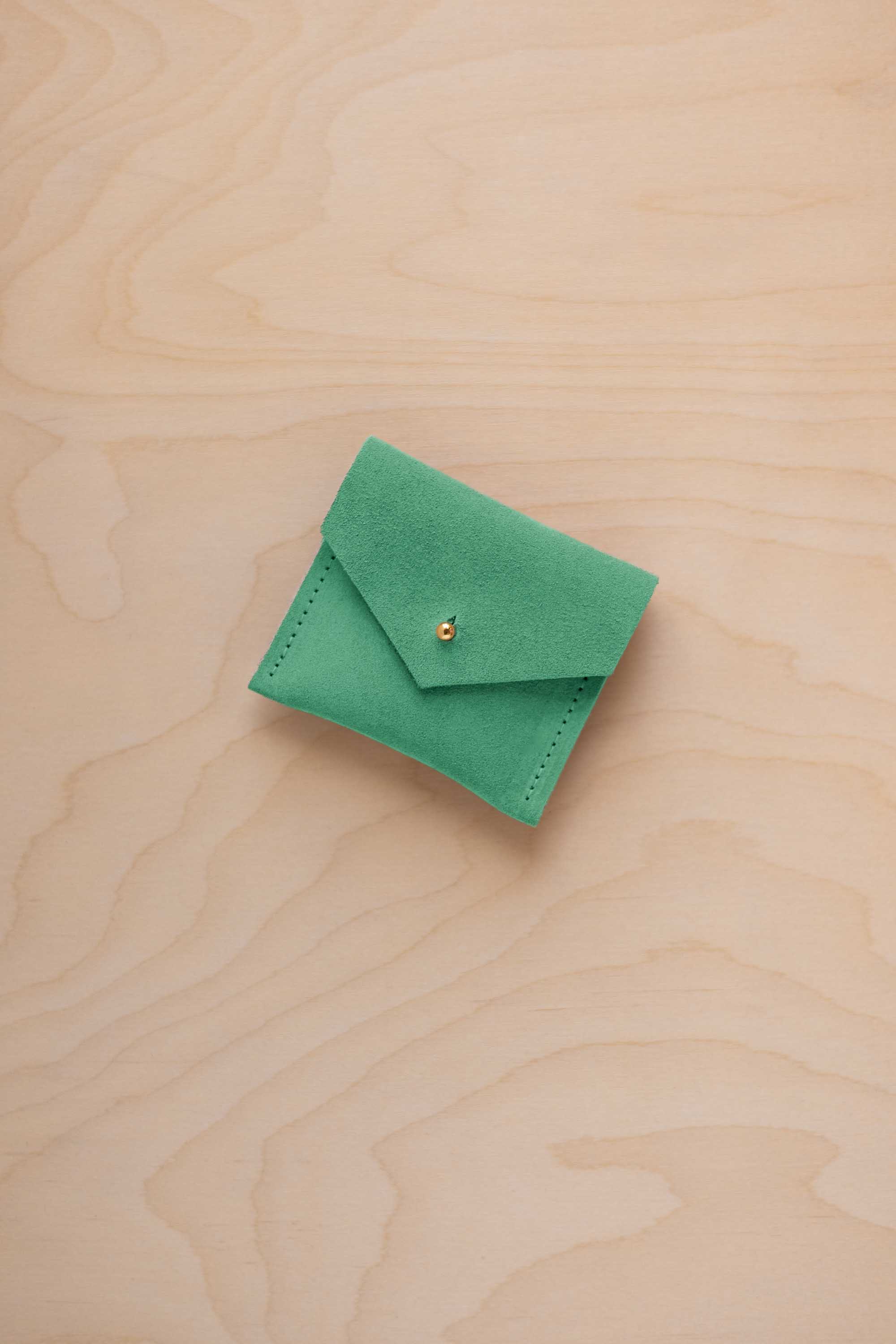 A small light emerald green suede pouch with a gold coloured button closure, placed on a light wood grain surface.