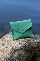 A small green suede envelope-style pouch with a gold button, resting on a large textured rock with a blurred outdoor background.