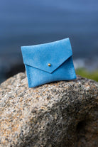 A small blue suede envelope-style pouch with a gold button, resting on a large textured rock with a blurred outdoor background.