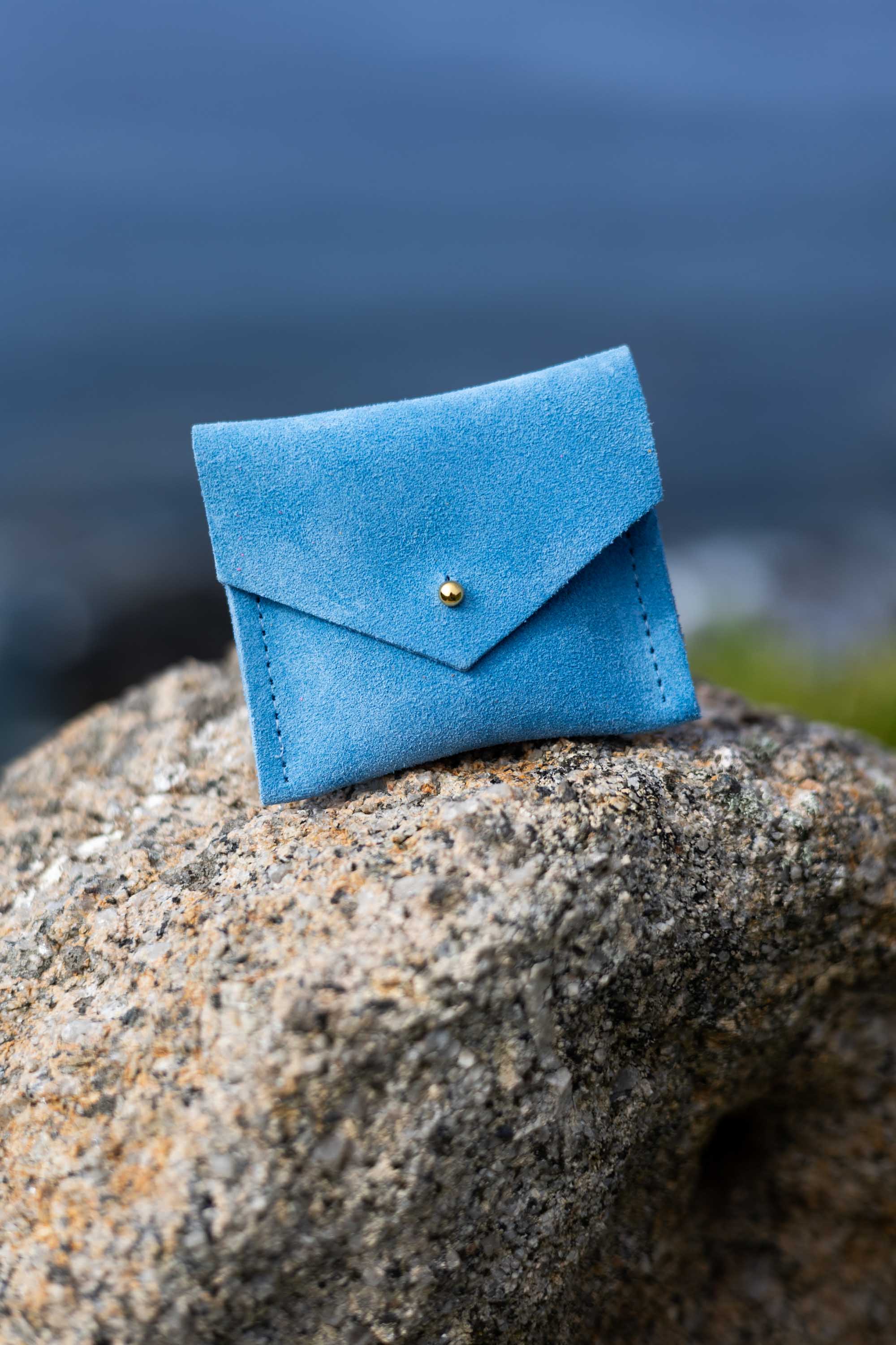 A small blue suede envelope-style pouch with a gold button, resting on a large textured rock with a blurred outdoor background.