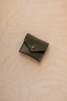 A small olive green suede pouch with a flap closure and gold button, placed on a light wood grain surface.