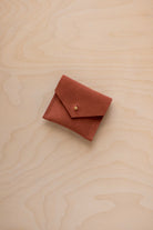 A small terracotta-colored suede pouch with a flap closure and a gold button, resting on a light wood grain surface.