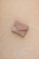 A small warm beige suede jewelry pouch with a gold button, resting on a light wood grain surface.