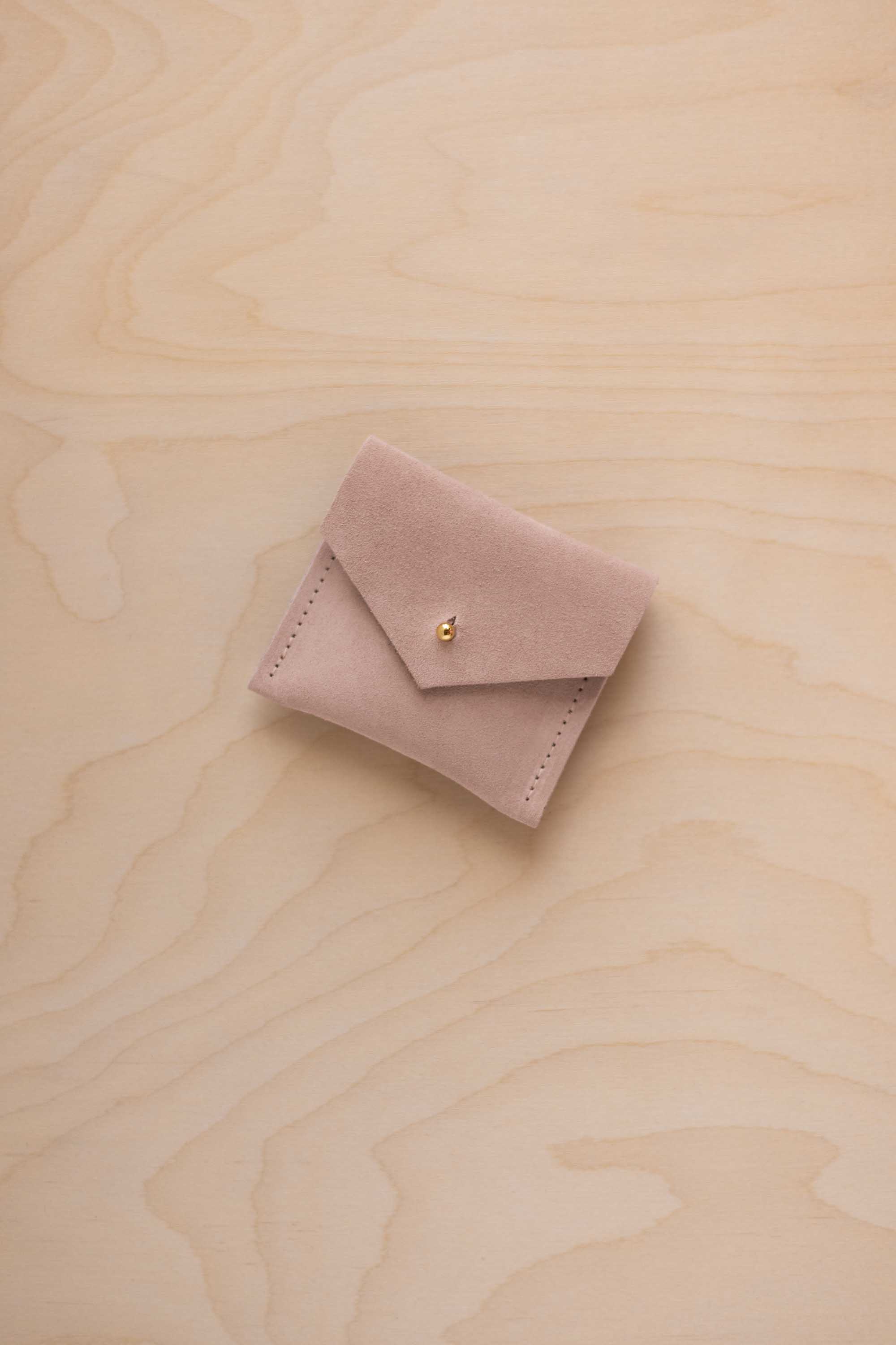 A small warm beige suede jewelry pouch with a gold button, resting on a light wood grain surface.
