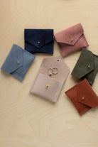 Six small suede envelope style pouches in different colours arranged on a light wood surface. The pouches are blue, navy, pink, olive green, blush, and rust, each with a small gold-tone stud closure.