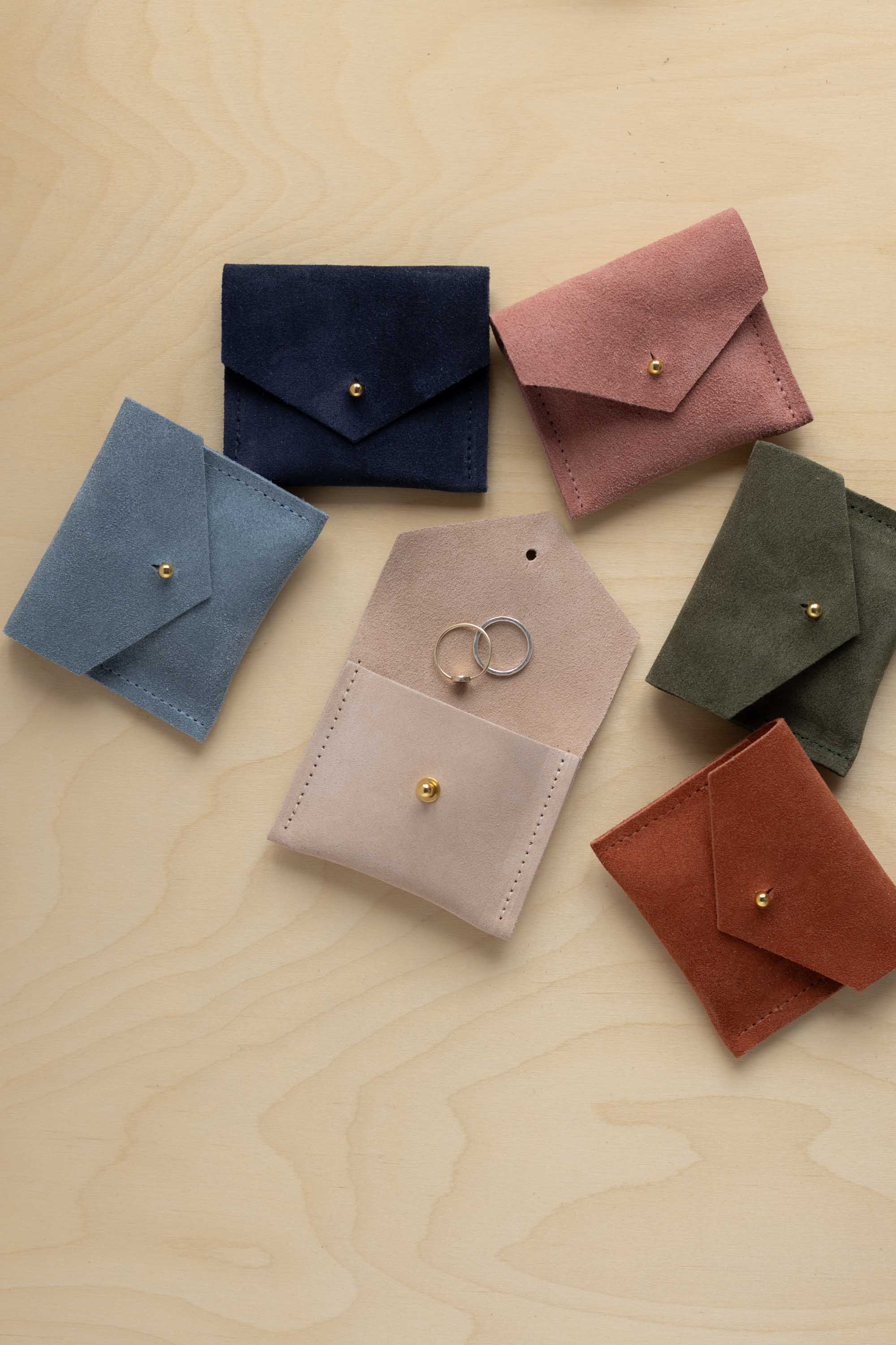 Six small suede envelope style pouches in different colours arranged on a light wood surface. The pouches are blue, navy, pink, olive green, blush, and rust, each with a small gold-tone stud closure.