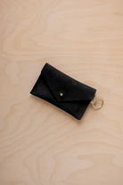 A small soft black coloured suede envelope-style keyring pouch with a gold button, resting on a light wood grain surface.
