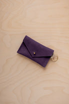 A small blackcurrant coloured suede envelope-style keyring pouch with a gold button, resting on a light wood grain surface.