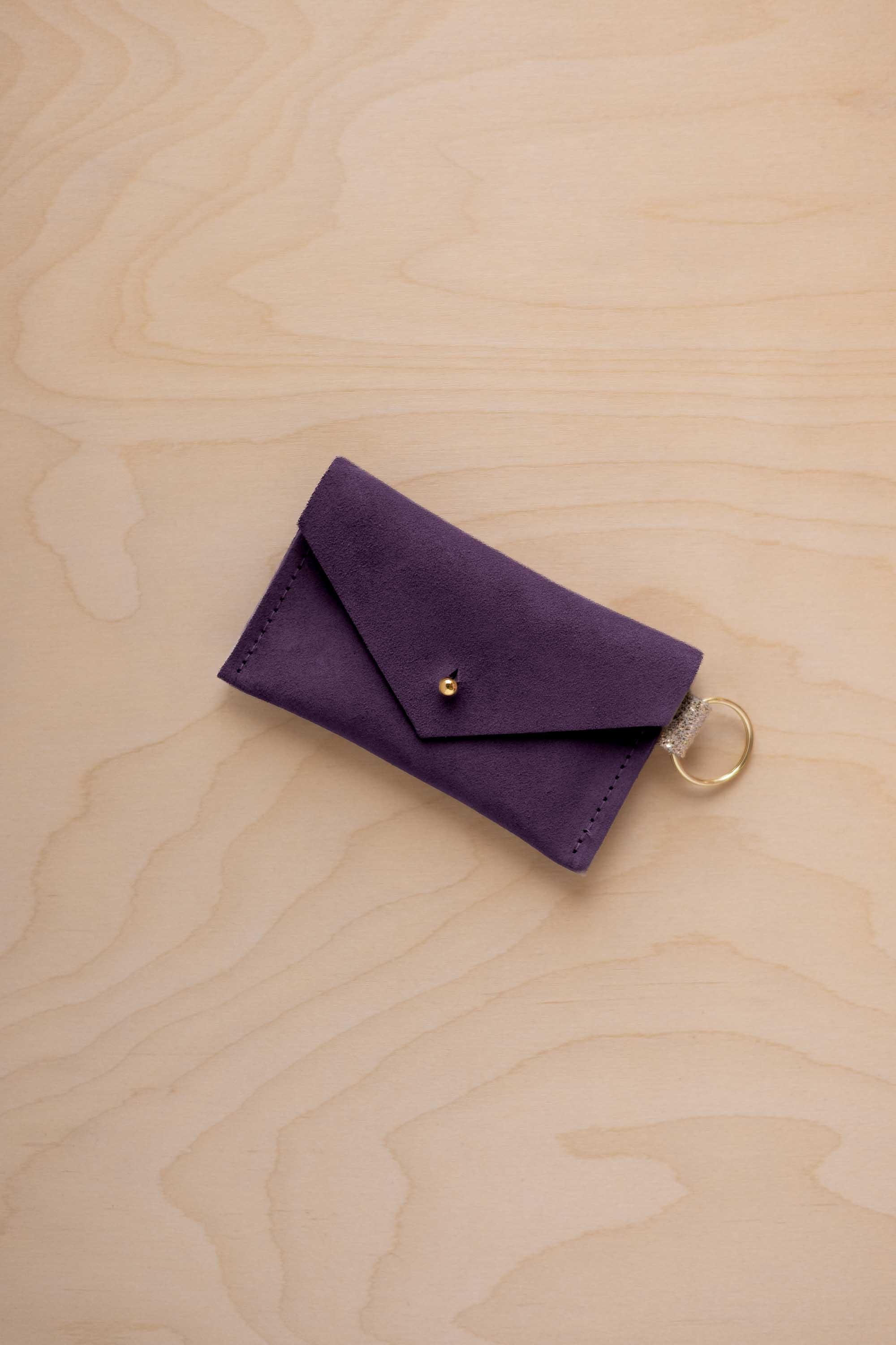 A small blackcurrant coloured suede envelope-style keyring pouch with a gold button, resting on a light wood grain surface.