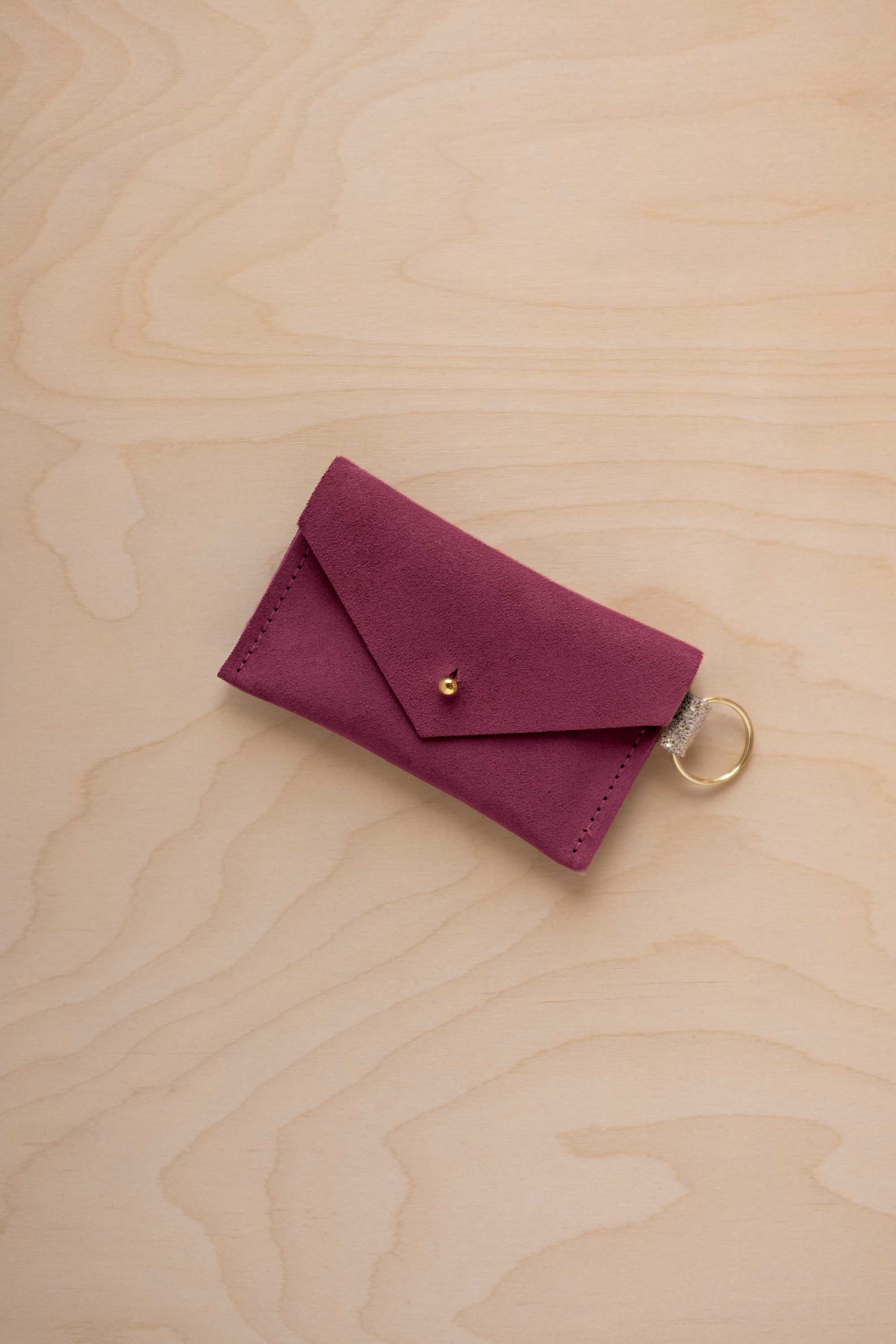 A small burgundy coloured suede envelope-style keyring pouch with a gold button, resting on a light wood grain surface.