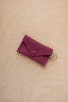 A small burgundy coloured suede envelope-style keyring pouch with a gold button, resting on a light wood grain surface.