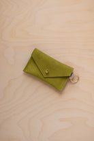 A small chartreuse green coloured suede envelope-style keyring pouch with a gold button, resting on a light wood grain surface.