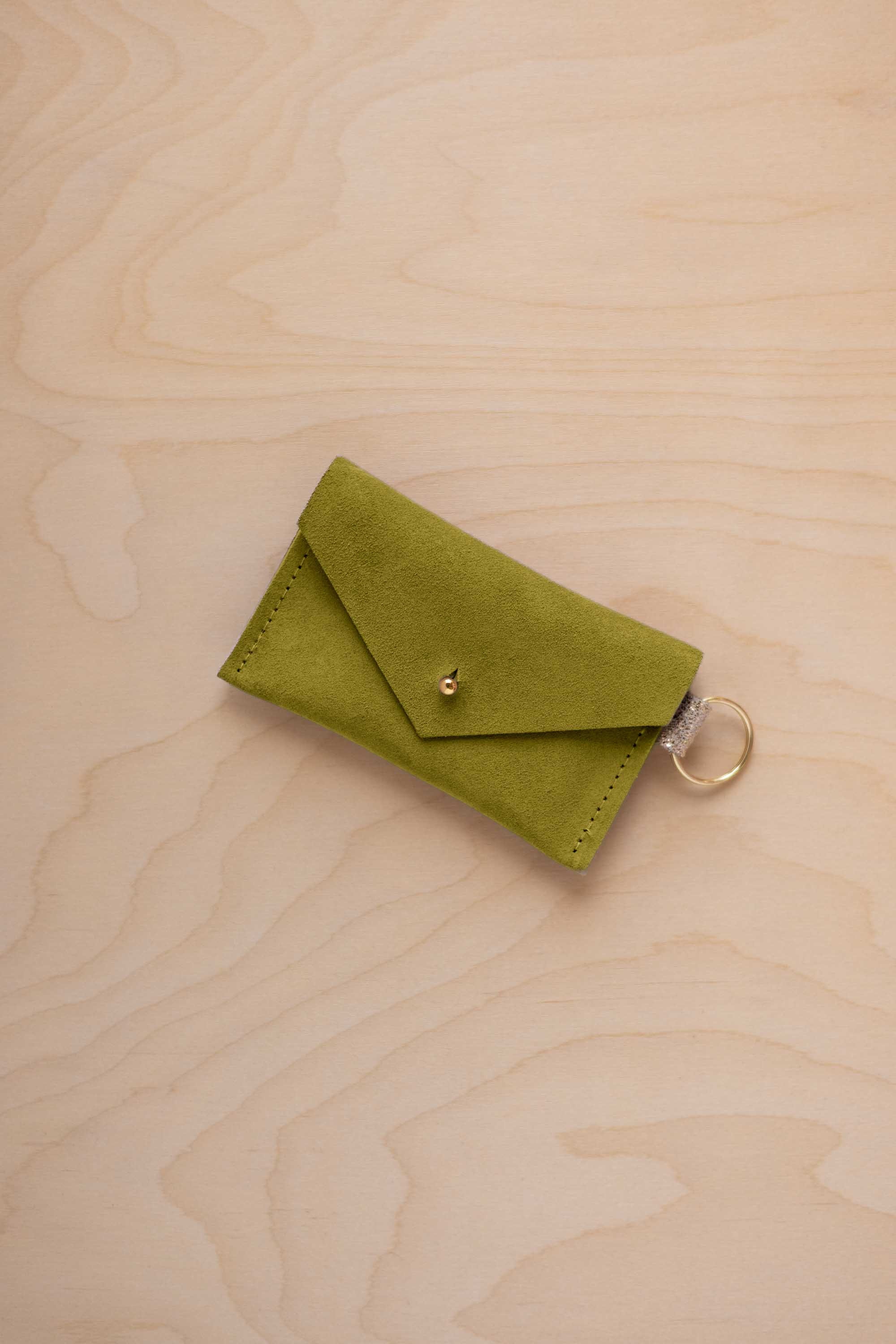 A small chartreuse green coloured suede envelope-style keyring pouch with a gold button, resting on a light wood grain surface.