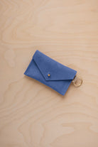 A small cobalt blue coloured suede envelope-style keyring pouch with a gold button, resting on a light wood grain surface.