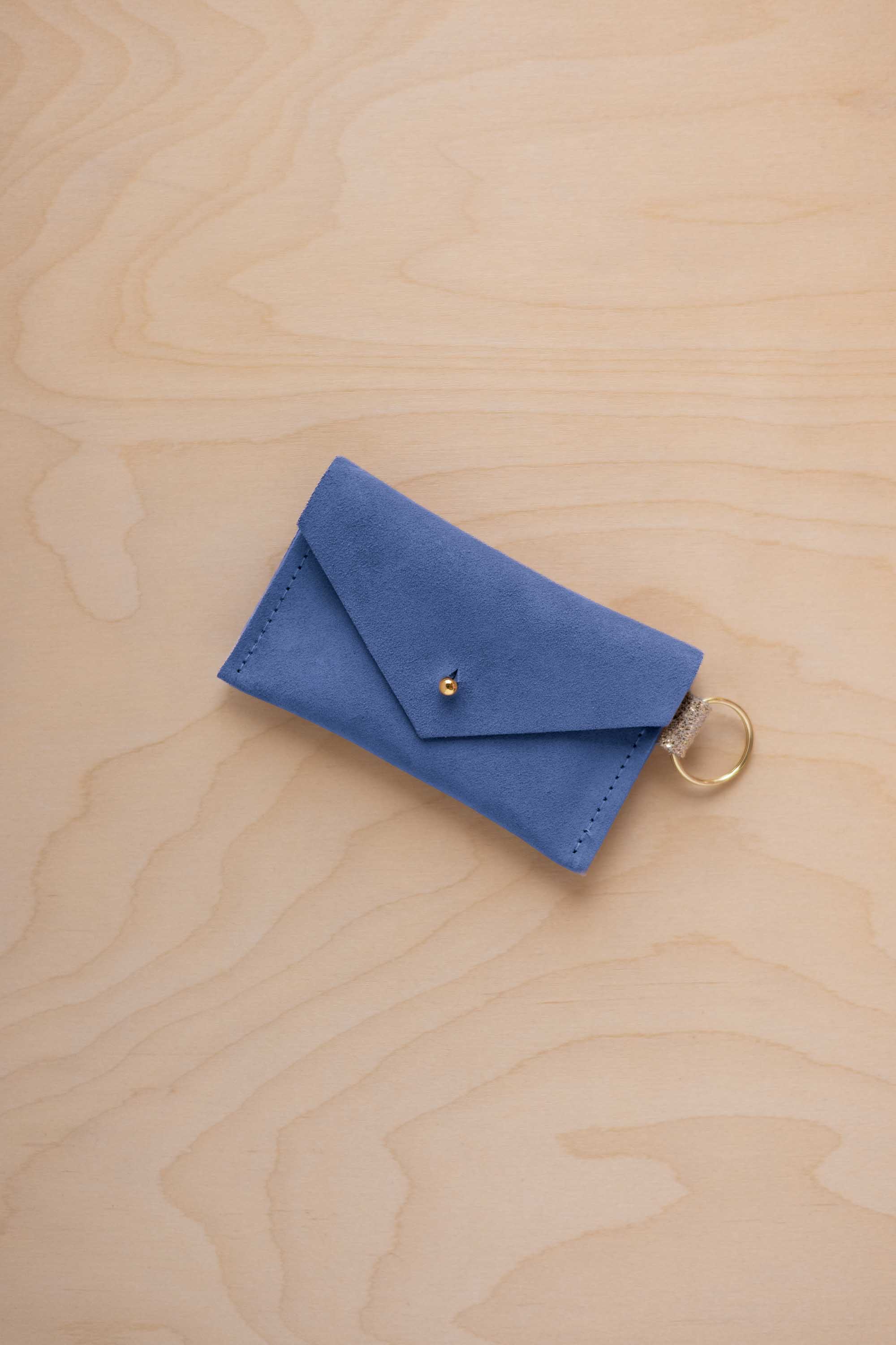 A small cobalt blue coloured suede envelope-style keyring pouch with a gold button, resting on a light wood grain surface.