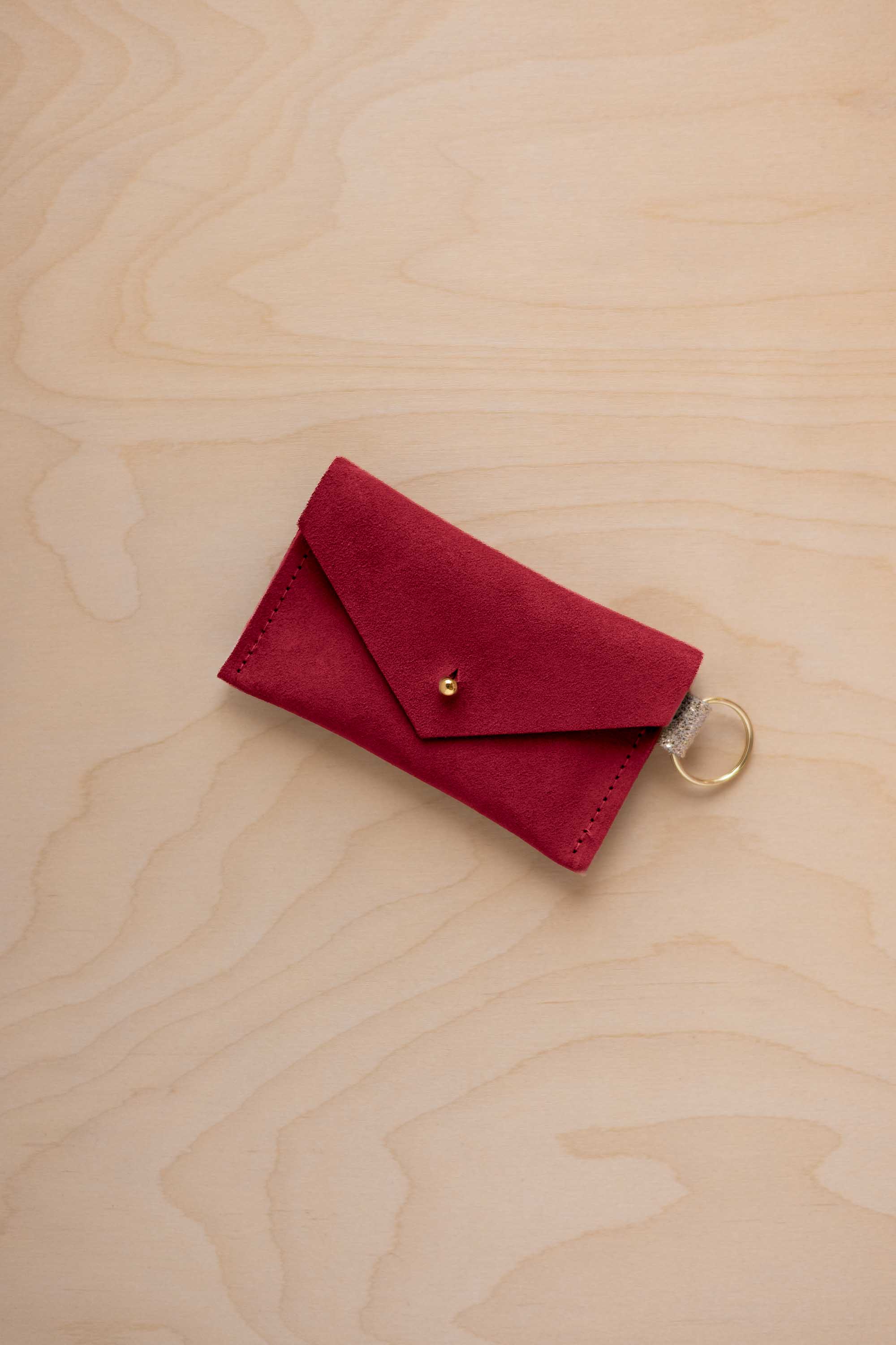 A small crimson red coloured suede envelope-style keyring pouch with a gold button, resting on a light wood grain surface.