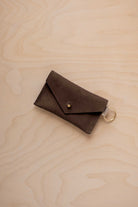 A small espresso brown coloured suede envelope-style keyring pouch with a gold button, resting on a light wood grain surface.
