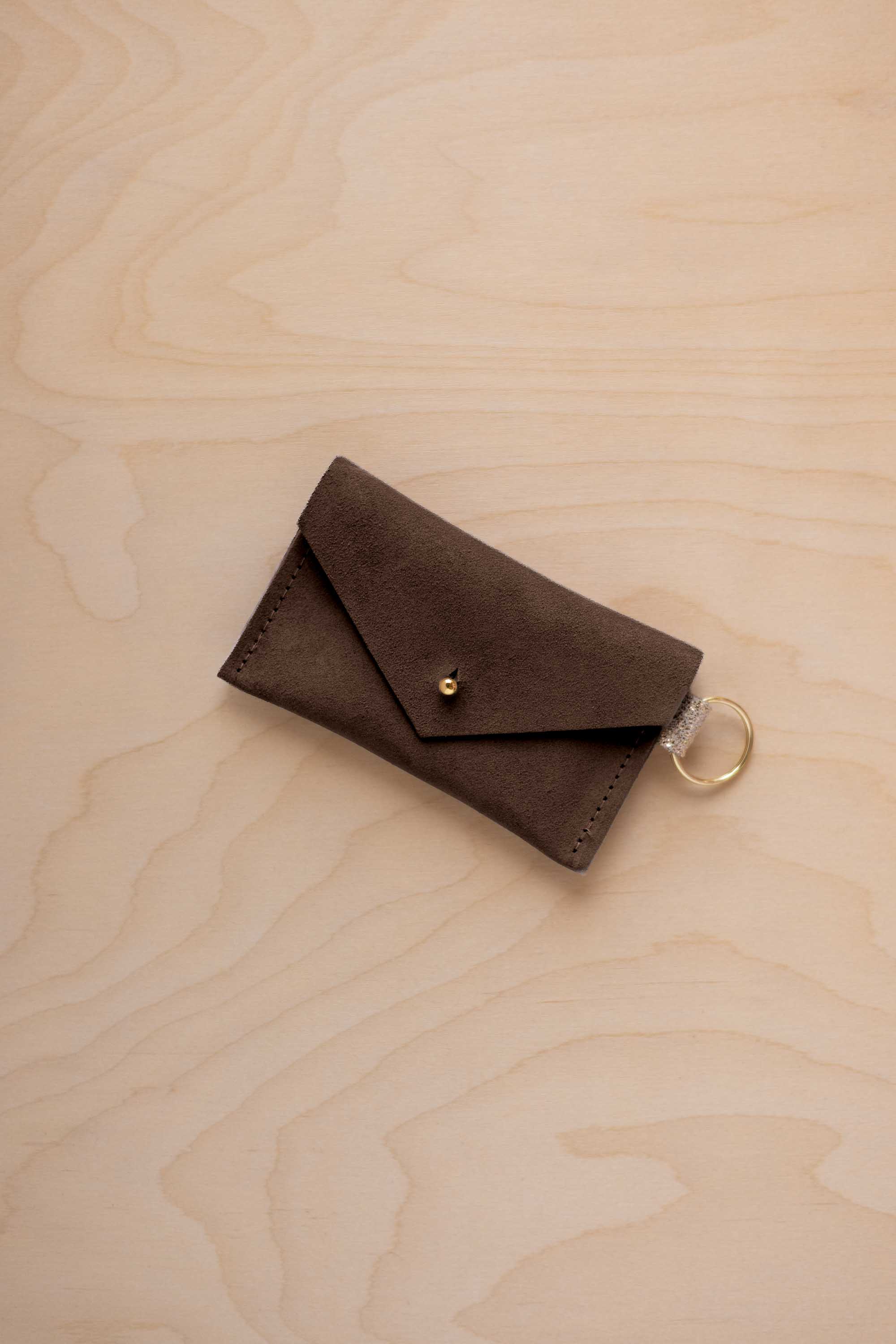 A small espresso brown coloured suede envelope-style keyring pouch with a gold button, resting on a light wood grain surface.