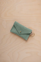 A small eucalyptus green coloured suede envelope-style keyring pouch with a gold button, resting on a light wood grain surface.