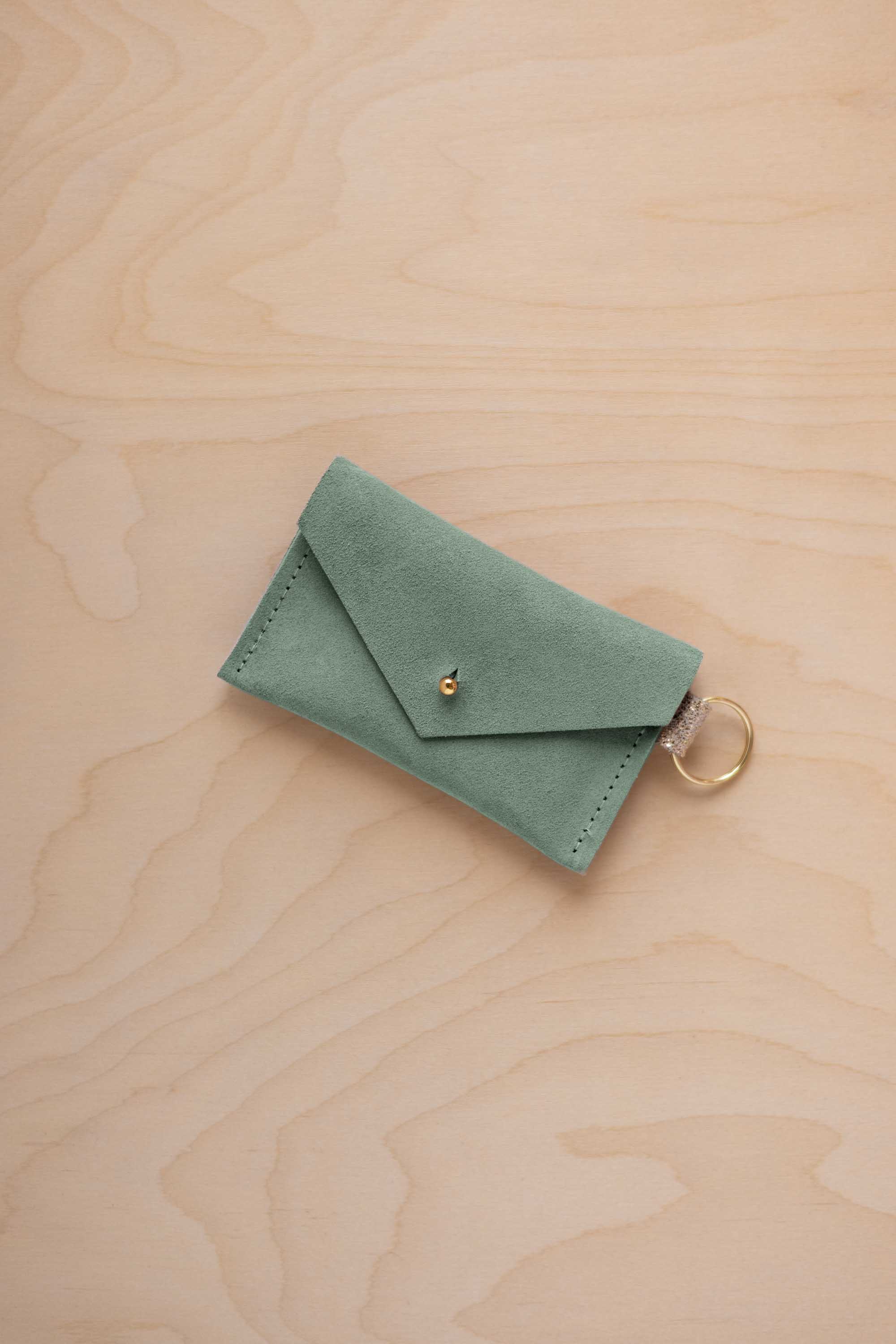 A small eucalyptus green coloured suede envelope-style keyring pouch with a gold button, resting on a light wood grain surface.