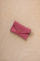A small faded rose coloured suede envelope-style keyring pouch with a gold button, resting on a light wood grain surface.