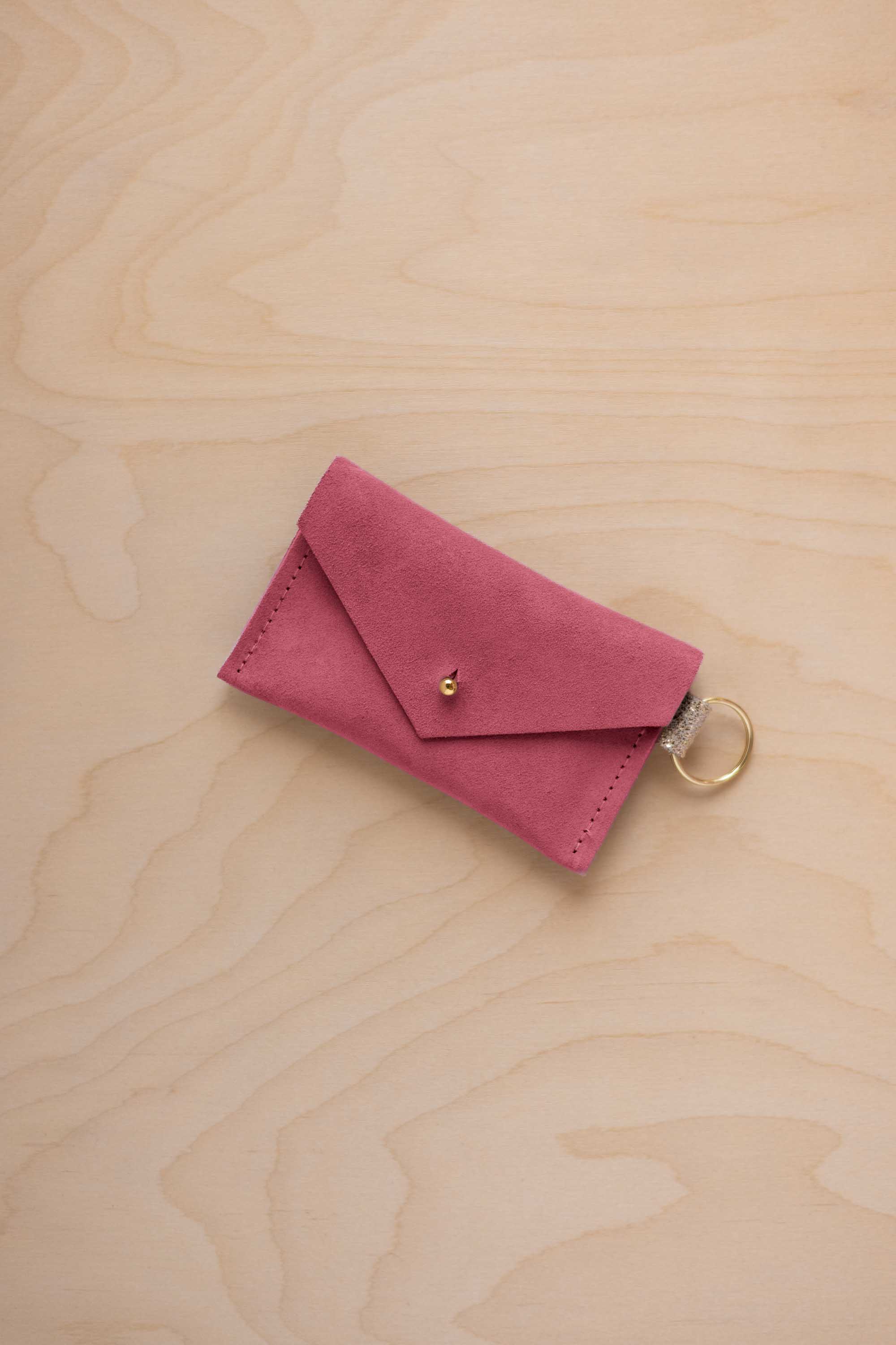 A small faded rose coloured suede envelope-style keyring pouch with a gold button, resting on a light wood grain surface.