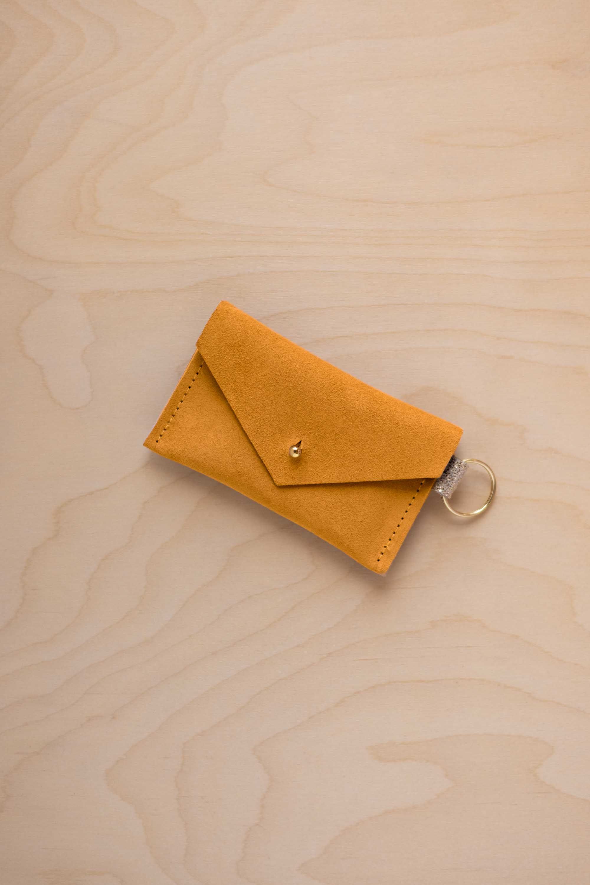 A small golden ochre coloured suede envelope-style keyring pouch with a gold button, resting on a light wood grain surface.