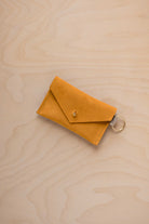 A small golden ochre coloured suede envelope-style keyring pouch with a gold button, resting on a light wood grain surface.