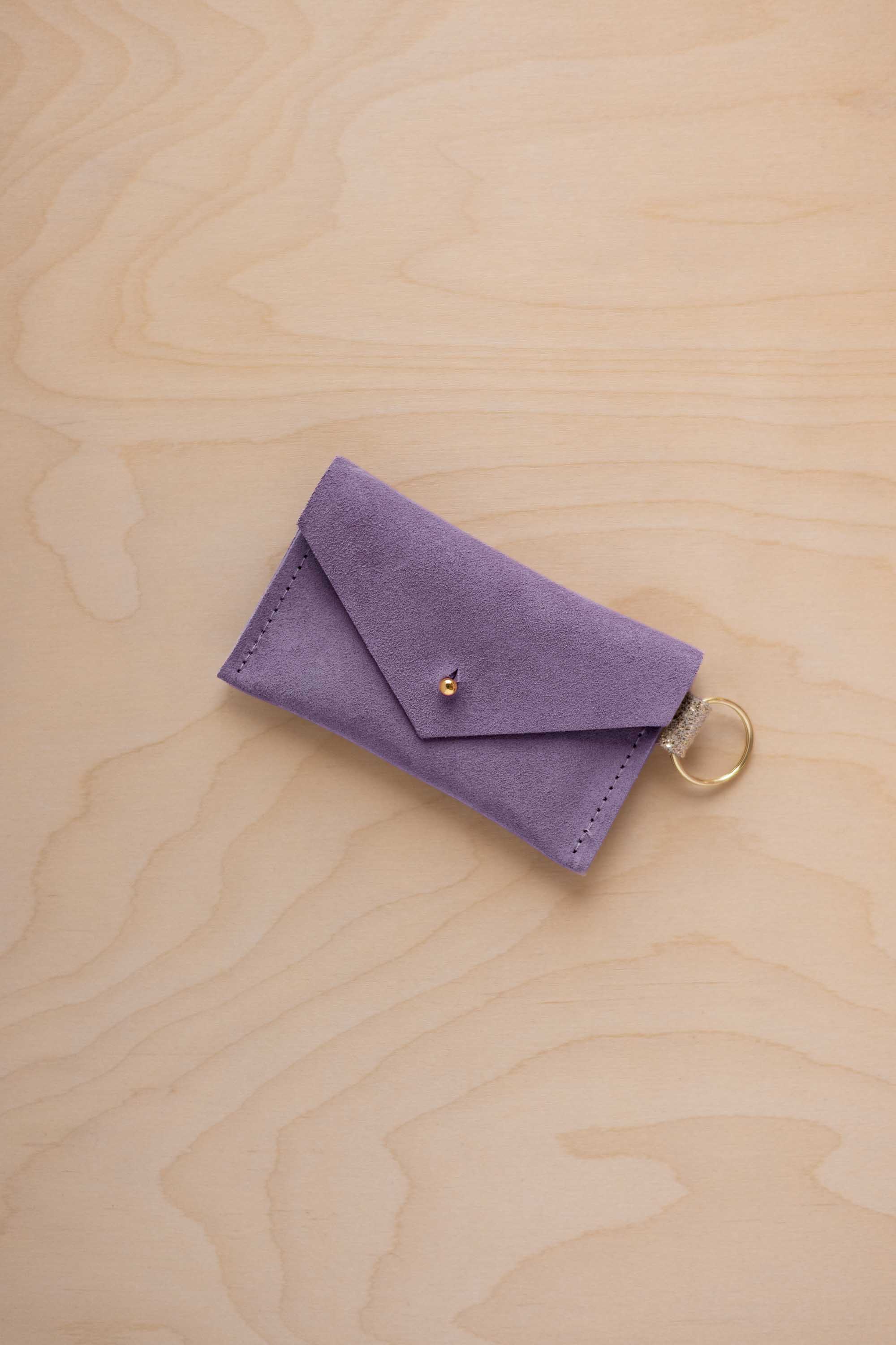 A small lavender coloured suede envelope-style keyring pouch with a gold button, resting on a light wood grain surface.